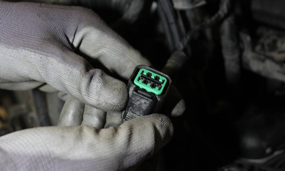 Gloved hands holding a rectangular four pin automotive connector as the mechanic gets ready to replace it inside of the car.