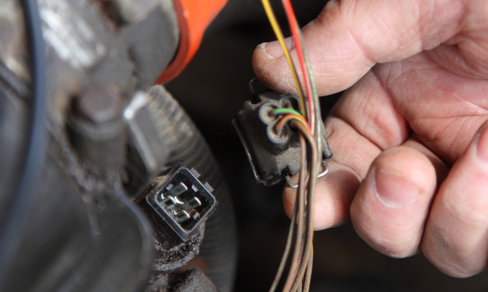 The hand of a mechanic holding a 4-pin automotive connector that&rsquo;s connected to the colored wires of the vehicle.
