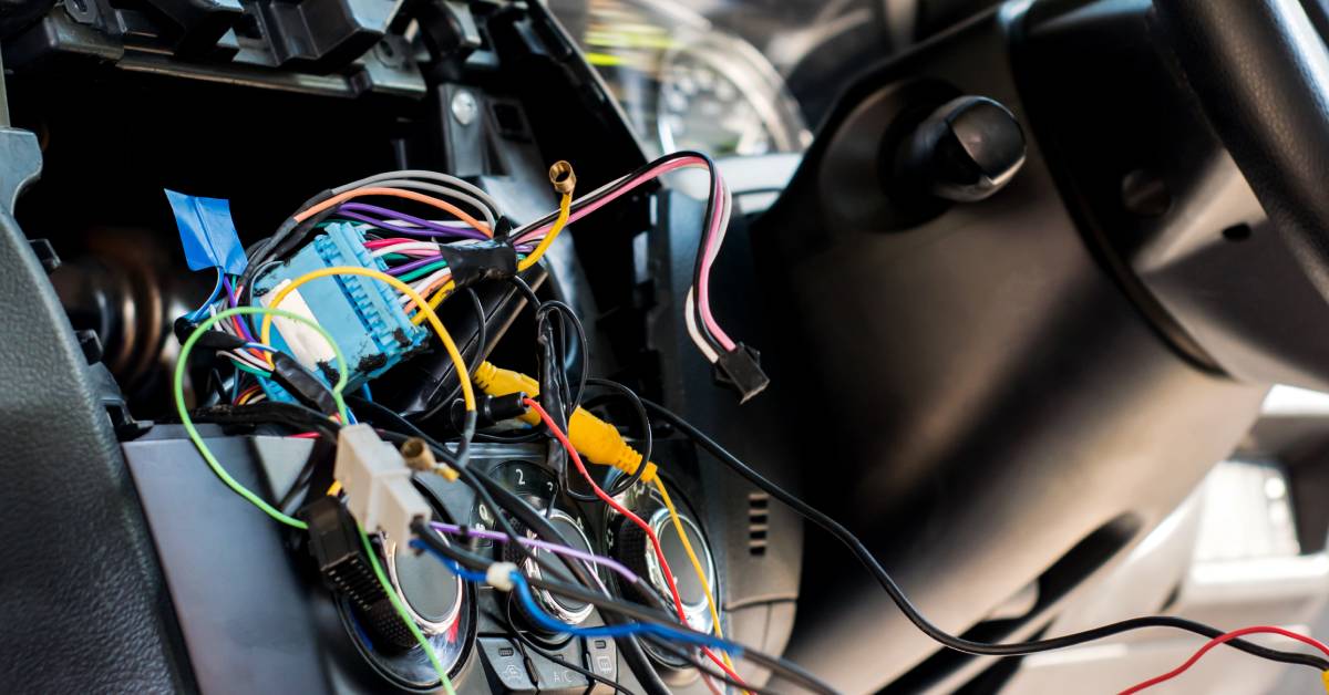The exposed wires and electric connectors of a car dashboard are beside the steering wheel and above the control panel.