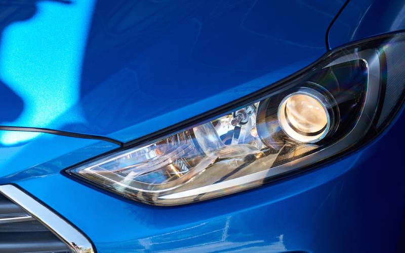  Closeup of the front headlight of a bright blue sedan. The circular lamp inside is shining brightly.