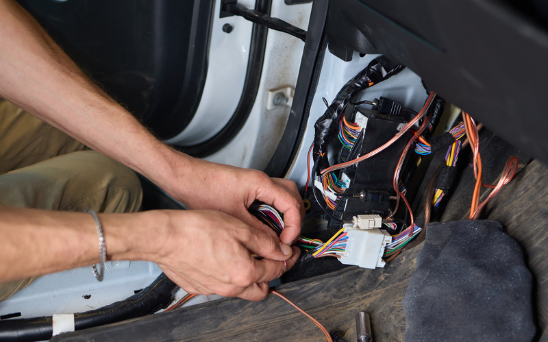 A close-up of a pair of hands working on a car&rsquo;s electrical system. The car is white and the wires are multicolored.