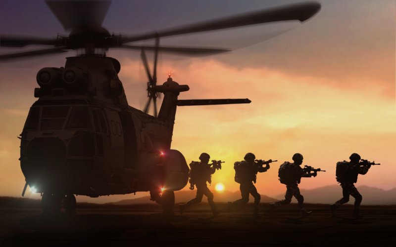 The silhouette of military members leaving a helicopter with their guns raised as the sun sets.