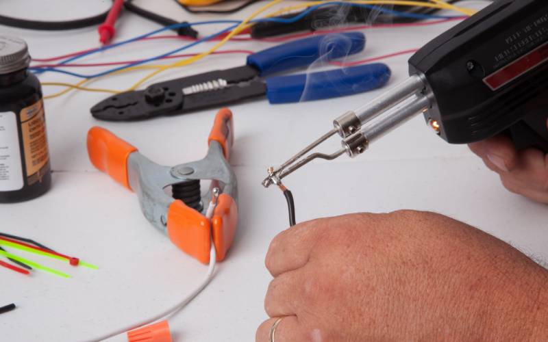 A close-up of a person using a soldering tool on a wire. The worktable is covered with other wires and tools.