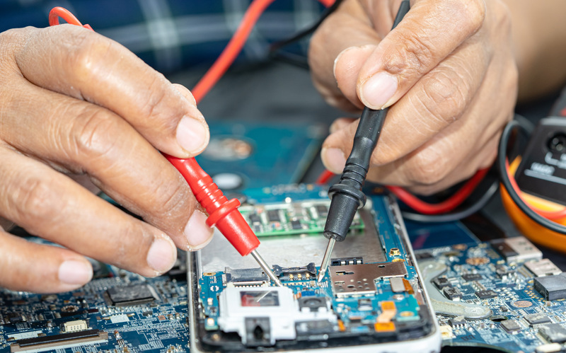 A pair of hands uses red and black tools to work on the internal electrical system of a smartphone on a table.