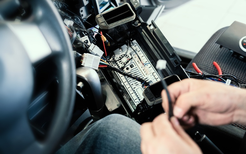The pair of hands holds a wire over a car radio that is turned upside-down revealing wires coming off of it.