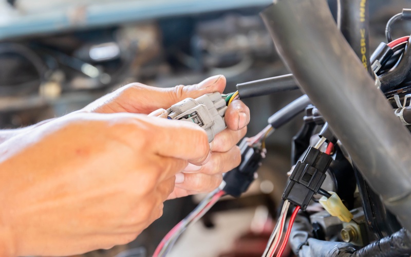 A pair of hands grips a gray plastic connector with colorful wires coming out of it attached to a car&rsquo;s electrical system.