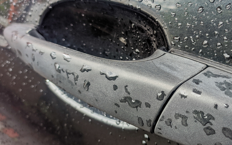A close-up of a car handle on a black car with rain drops on it and the body of the car.