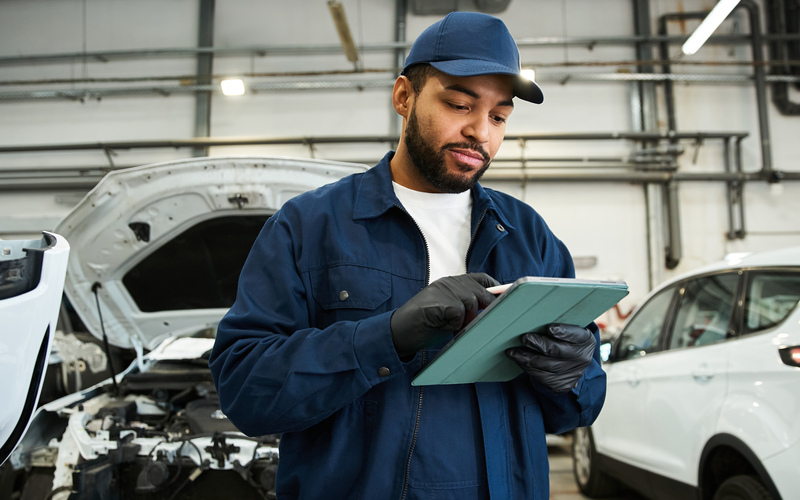 A man wearing a blue jacket and baseball cap looks at a tablet in an automotive garage.