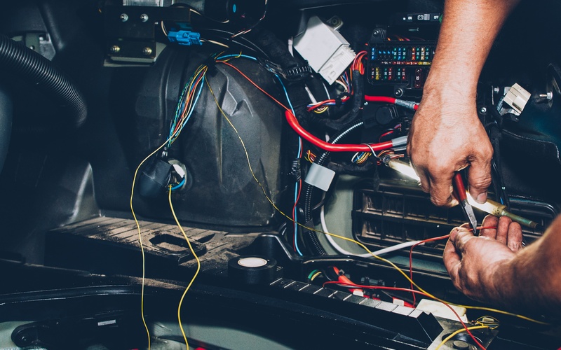 A pair of hands uses a tool on an orange wire that is part of a vehicle&rsquo;s electrical system.