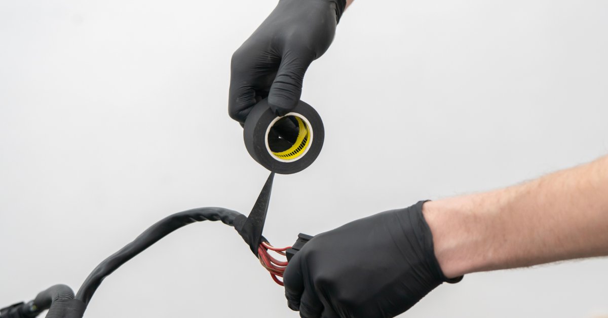 A pair of hands wearing black latex gloves wraps black electrical tape around multiple red wires.