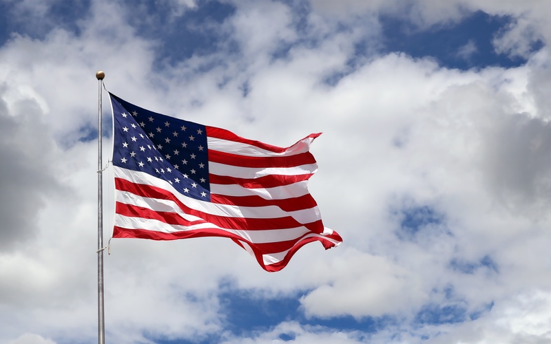 An American flag on a flagpole blows in the wind in front of a blue sky with some white clouds.