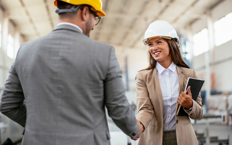A man in a gray suit and yellow hard hat shakes hands with a smiling woman in a beige suit and white hard hat.