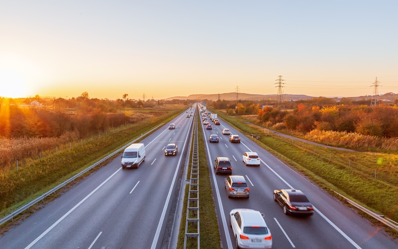 A two-lane highway near a mountain with cars driving in both directions. In the background, the sun is setting.