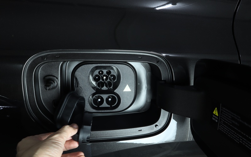 A person using their hand to open a panel on the exterior of an electric vehicle. The panel contains a 9-pin outlet.