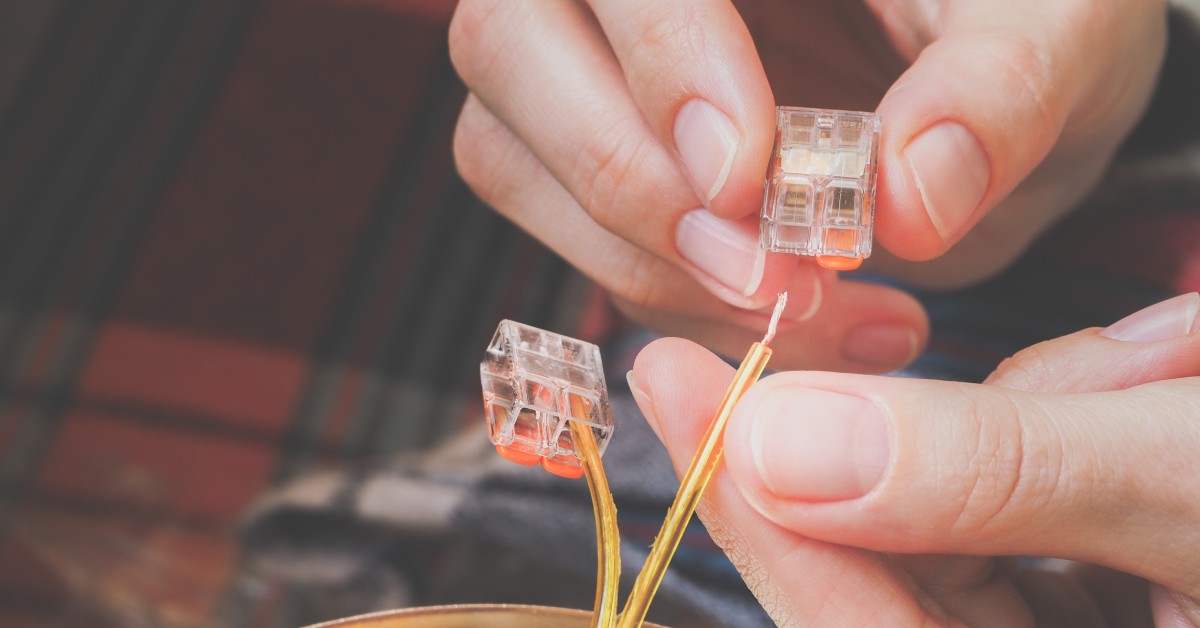 A pair of hands is pinching thin, gold wiring and inserting it into a clear 2-pin electrical connector.