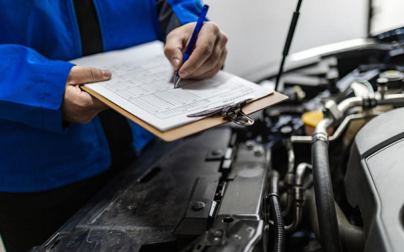 A person wearing a bright blue jacket uses a pen to write on a clipboard next to the open hood a car.