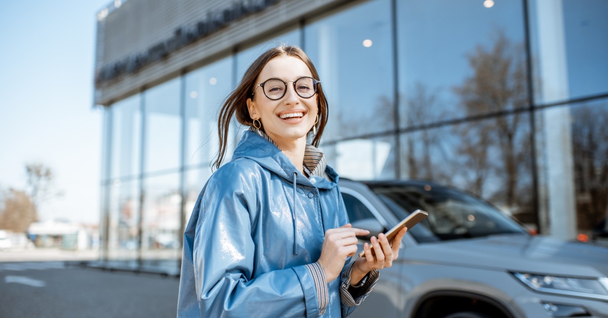 A person wearing glasses and a light blue hoodie standing next to a gray car outside during the daytime.
