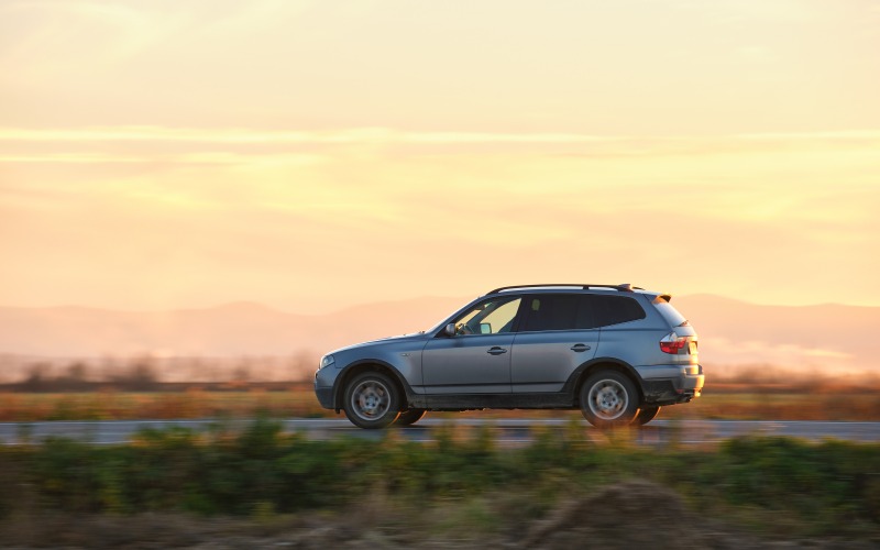 A small gray car driving alone near mountains on a road. The sunset turns the sky above the car orange.