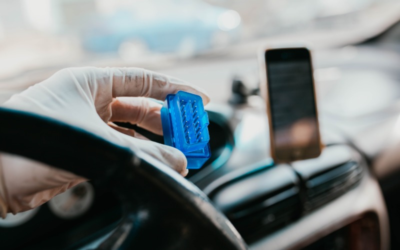 A person wearing a white glove holding a blue automotive connector in front of the steering wheel in their car.