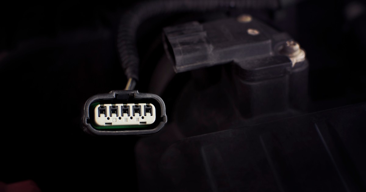 A black automotive five-pin connector with a white socket. The connector is attached to a car engine.