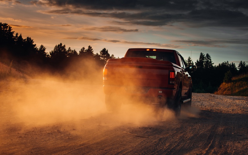 A truck driving down a dusty dirt road under an overcast sky, The truck is kicking up dirt from the road in its wake.