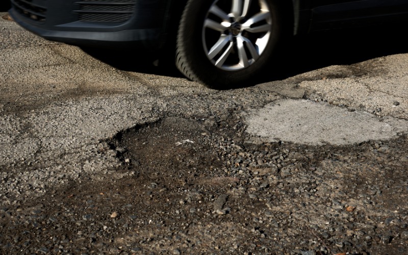A black car driving on a damaged road during the daytime. A huge pothole is in the middle of the road.