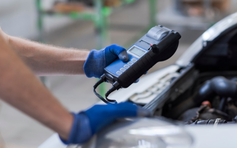 A mechanic wearing blue gloves holding a large black battery testing device over the open hood of a car. 