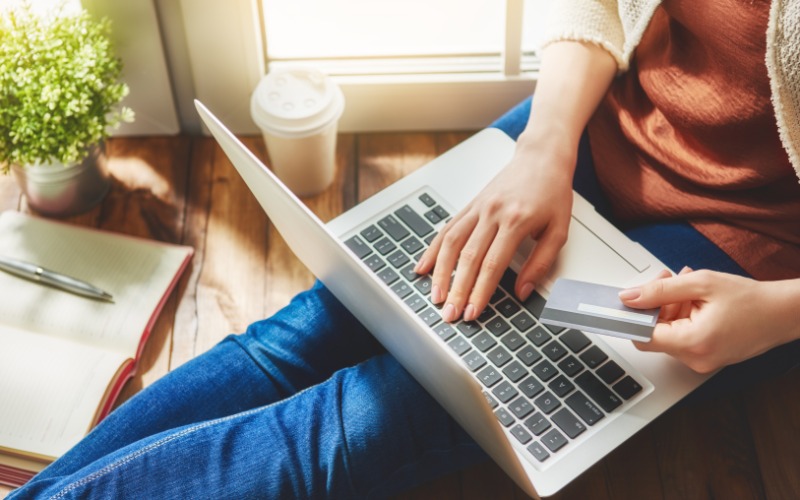 A person sitting on a table near a window holding a credit card. The person types on a laptop with their right hand.