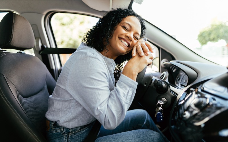 A person smiling and leaning on the steering wheel in their car. The car is parked outside during the daytime.
