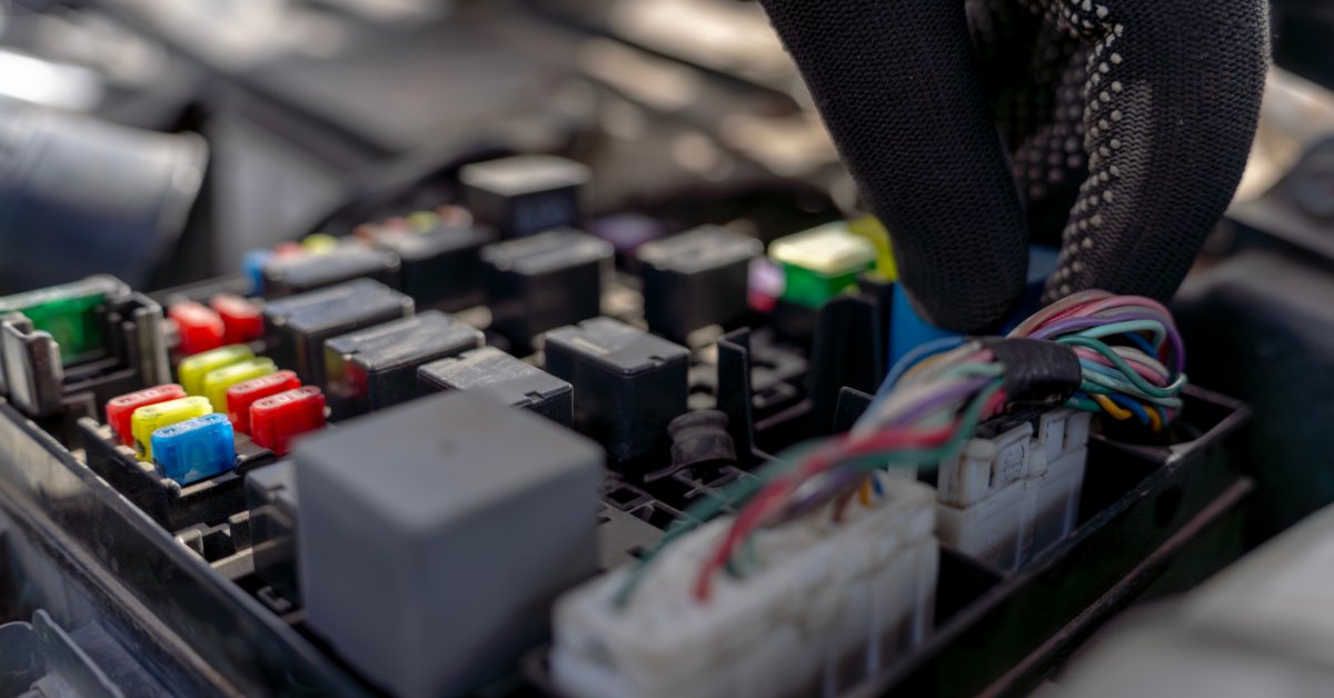 A mechanic wearing black gloves handling an automotive electrical connector installed on a car fuse relay block.