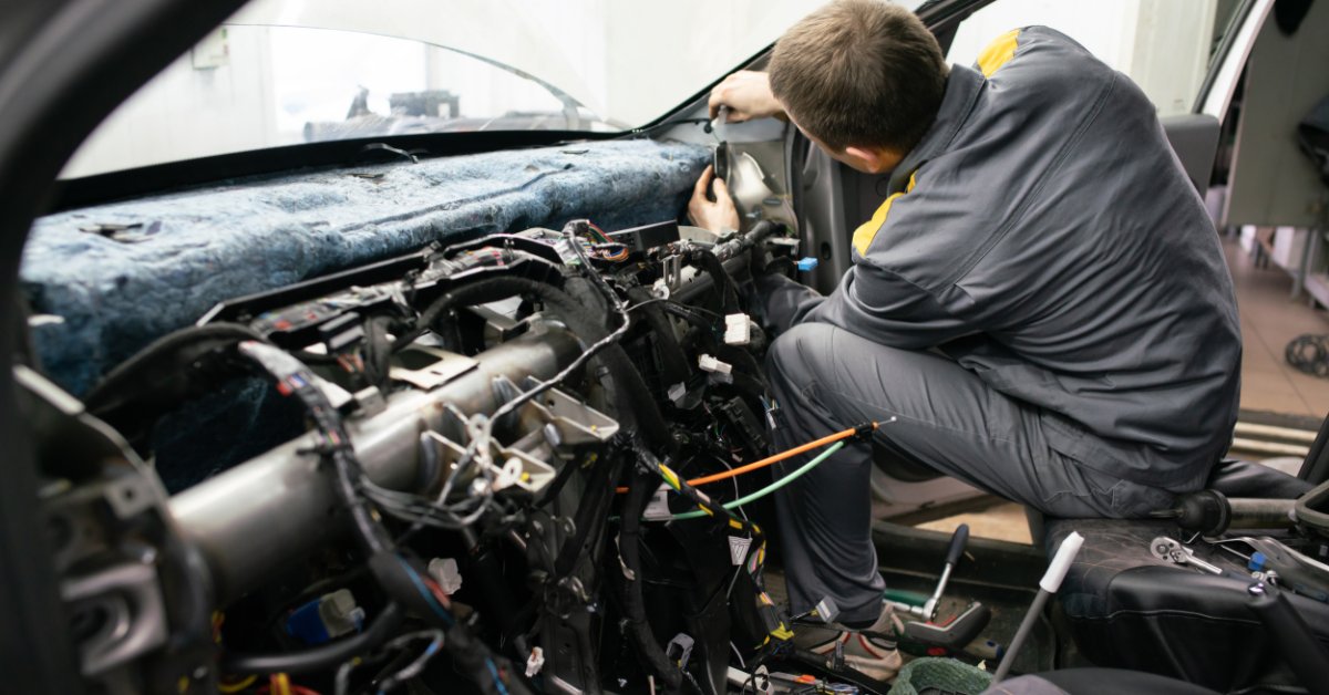 A mechanic wearing a gray uniform working on an exposed dashboard inside a car parked in an auto repair shop.