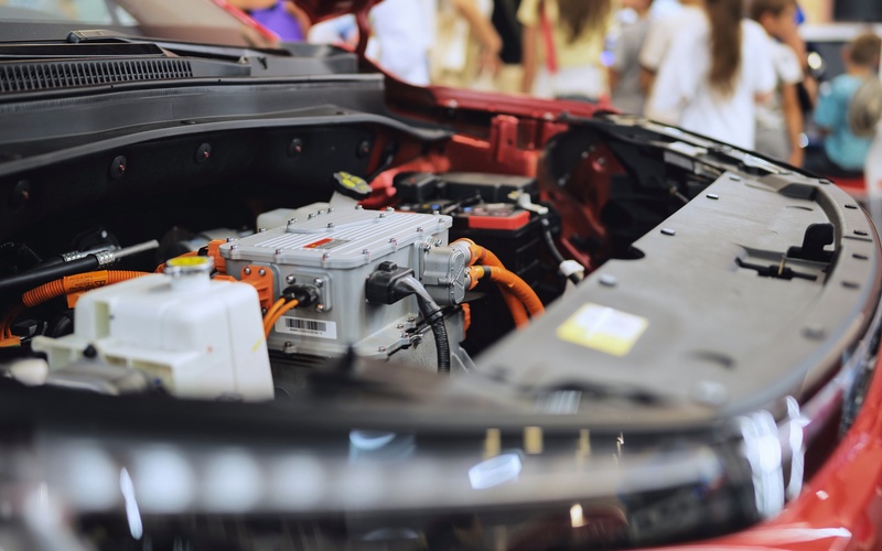 A red car parked in a brightly lit room. The engine bay is open to reveal the engine, wires, and connectors inside.