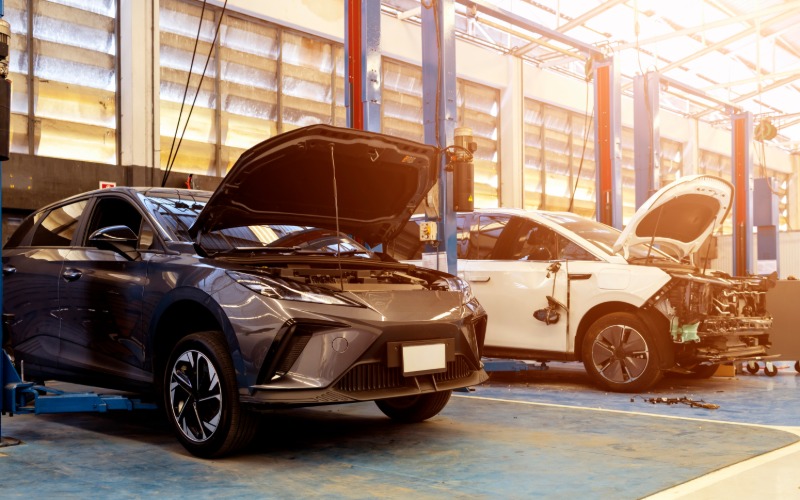 A black car and white car parked inside a repair shop during the daytime. Both car hoods are raised open.