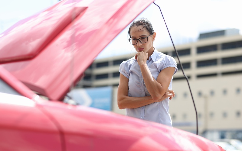 A person wearing glasses standing in front of a bright red car outside. The car&rsquo;s hood is propped open.