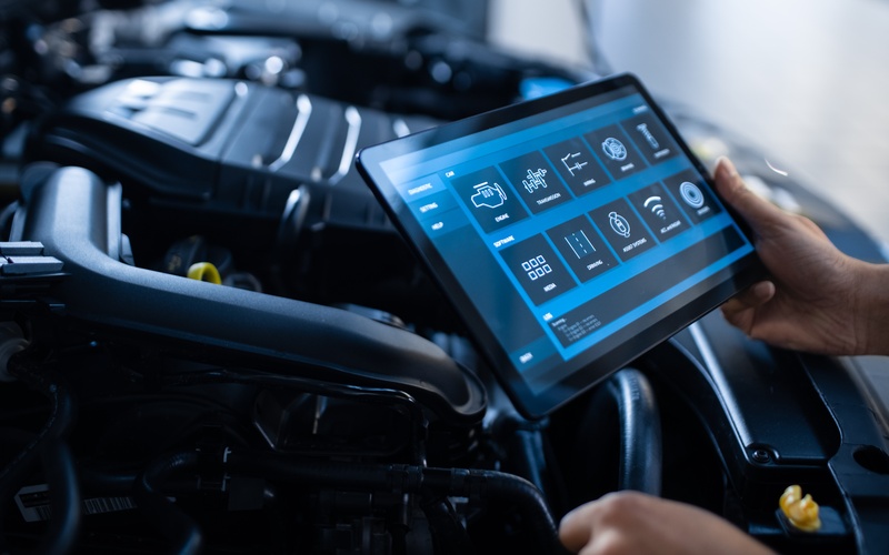 A close-up of a person holding a tablet with icons representing different parts of a car next to a car&rsquo;s engine.