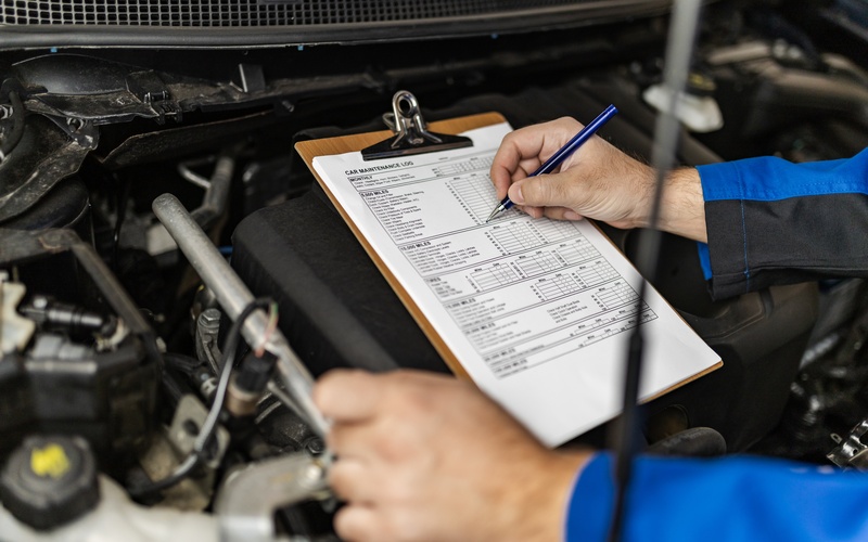 A close-up of a person holding a pen to a car maintenance log on a clipboard that is set on top of a car engine.