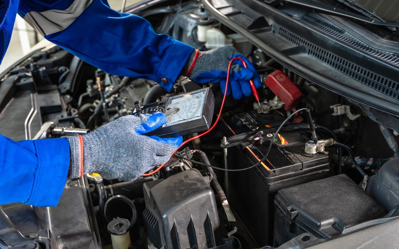 A close-up of a mechanic in a blue shirt and gray gloves holding a voltmeter to a car&rsquo;s battery.