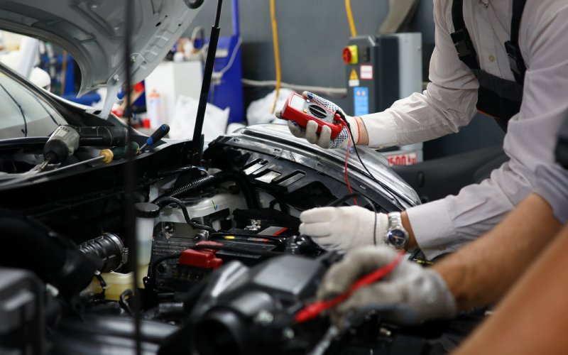 Two people wearing work gloves while using various devices to inspect the electrical system of a car.