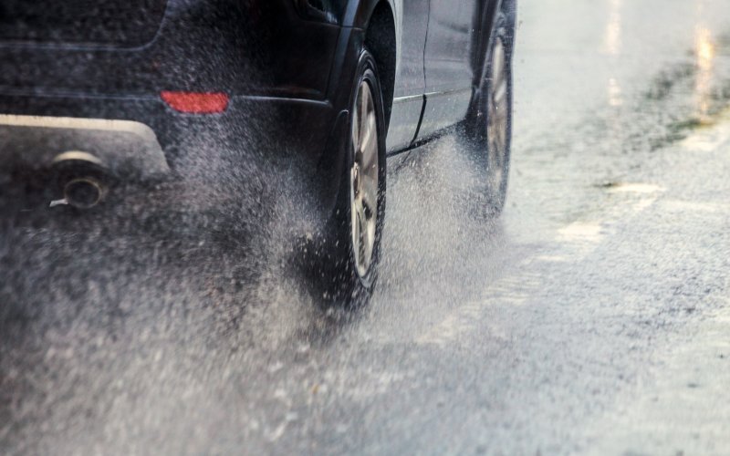 A close-up of a black car driving down a wet road, kicking up water behind its fast-spinning wheels.