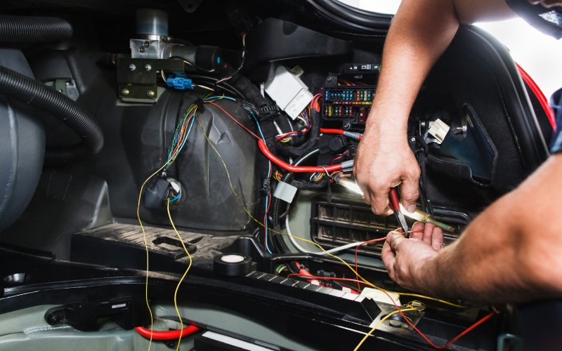 A close-up of a person using a red tool on an orange wire that is part of a vehicle&rsquo;s electrical system.